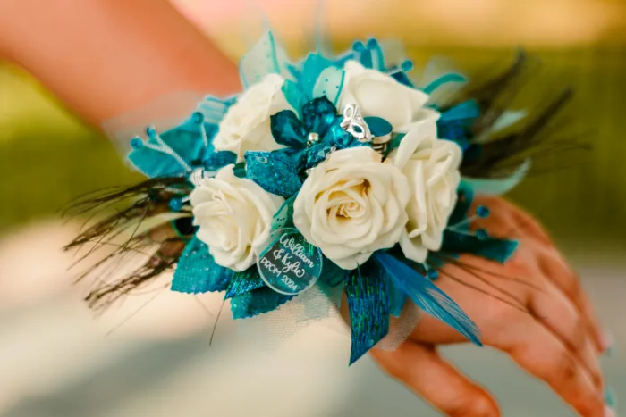 Close-up of a teal corsage with white roses, a personalized charm, peacock feathers, and a small masquerade mask pin for Prom 2024.