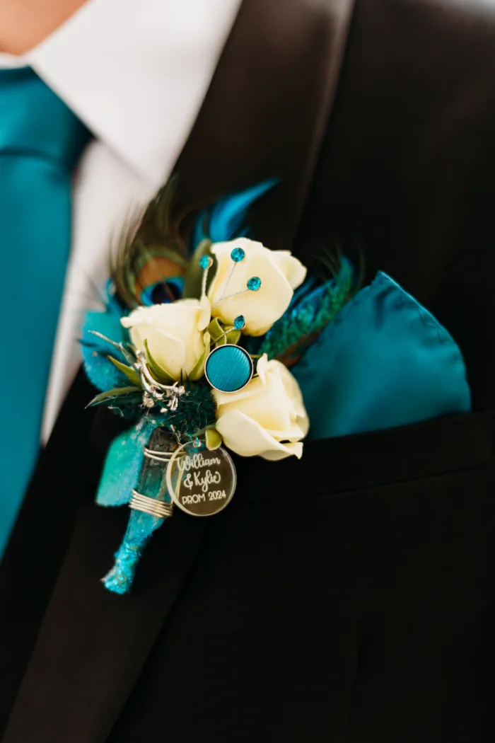 Close-up of a personalized teal boutonniere featuring peacock feathers, white roses, and a charm with 'William & Kylie Prom 2024' engraved.