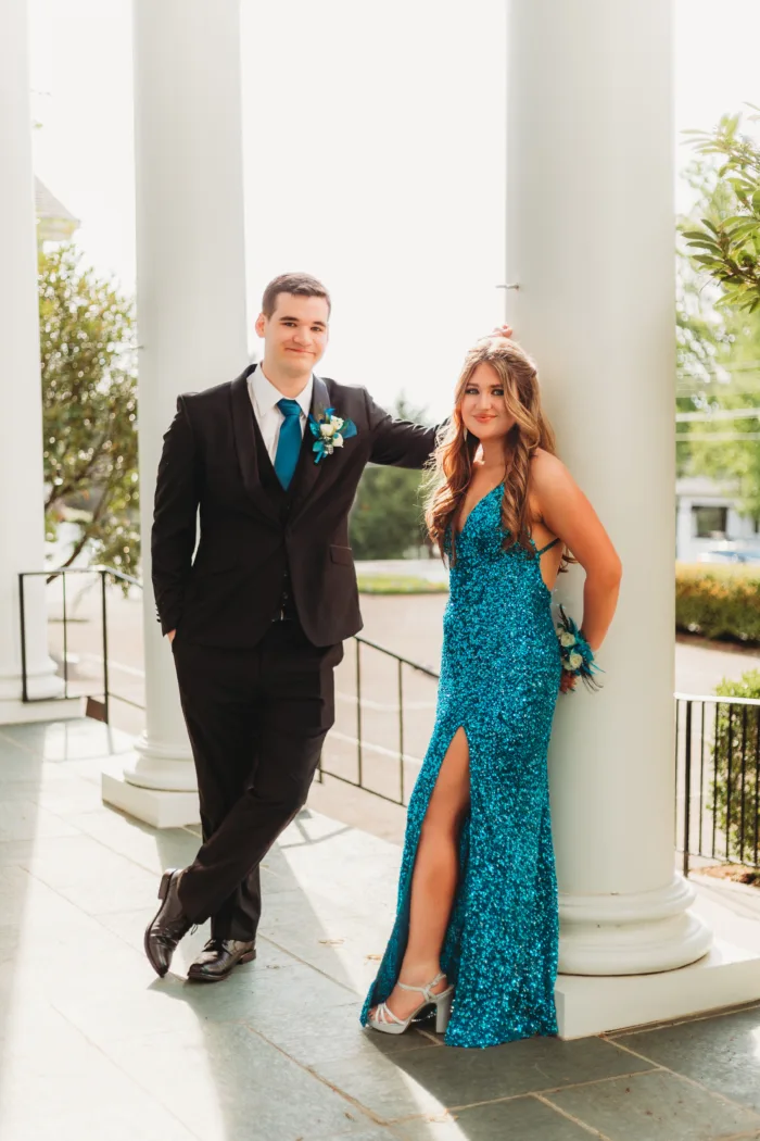 A young couple poses for Prom 2024, with the young woman in a teal sequin dress leaning against a column and the young man in a black tuxedo.