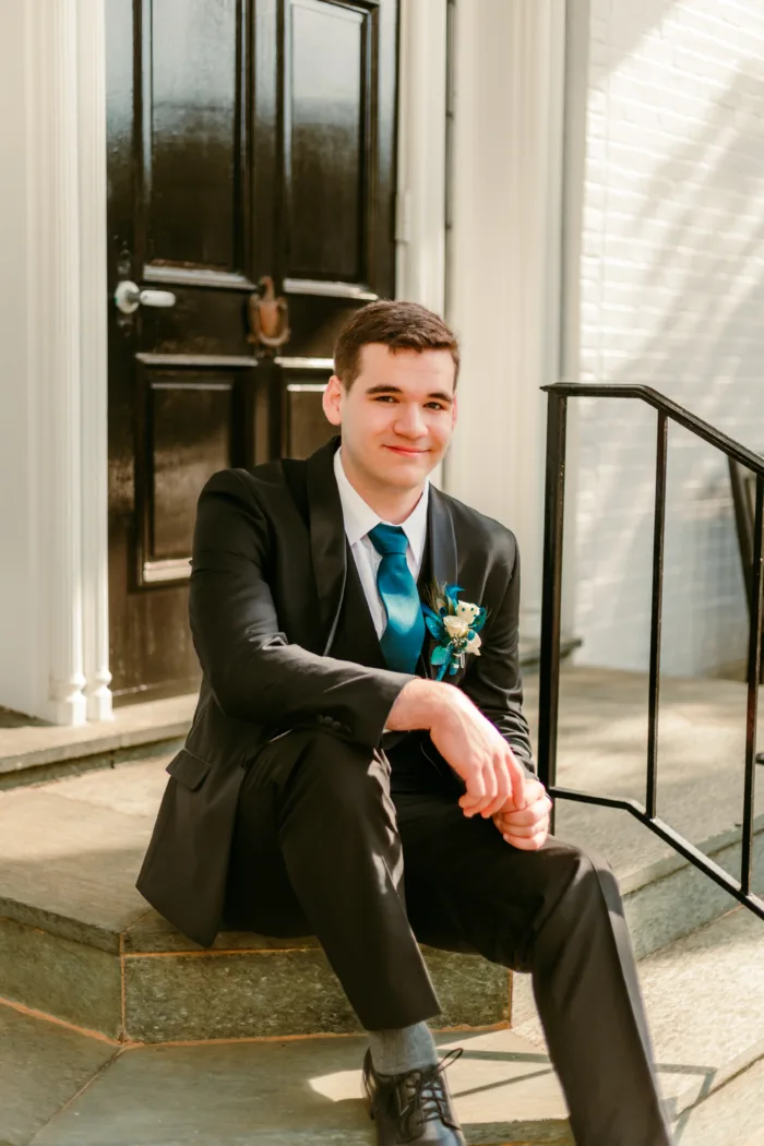Young man sitting in a black tuxedo with a teal tie and boutonniere, ready for Prom 2024.