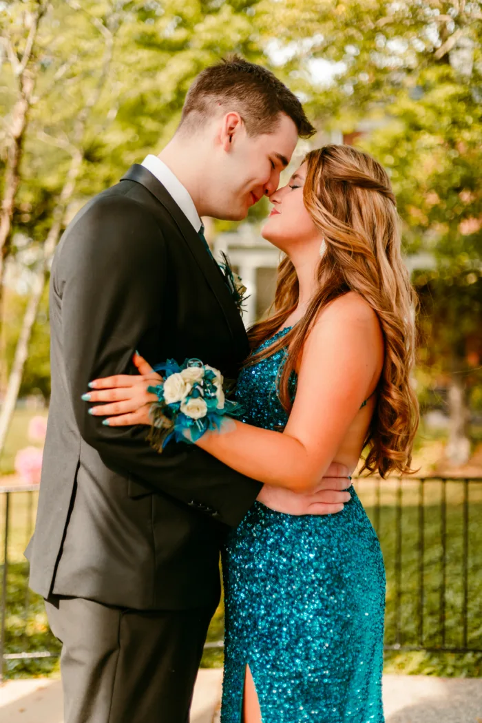 A young couple embraces before Prom 2024, with the young woman in a teal sequin dress and the young man in a black tuxedo.