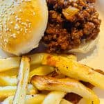 A homemade sesame seed hamburger bun filled with sloppy joe meat, served with seasoned fries on a plate.