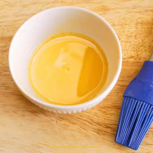 Egg wash in a white bowl next to a blue silicone pastry brush, perfect for achieving a golden finish.