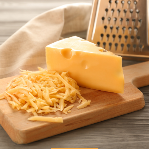 Block of cheddar cheese being freshly grated on a wooden board with a stainless steel grater.