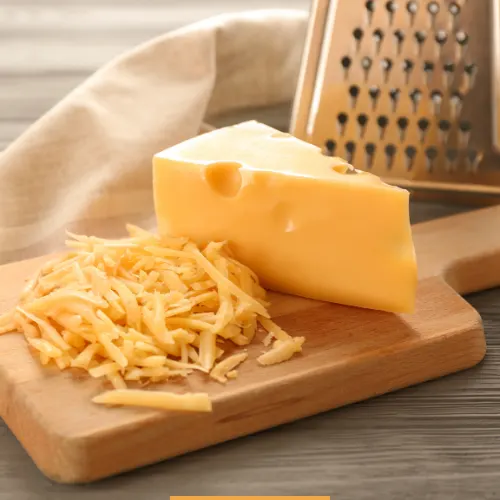 Block of cheddar cheese being freshly grated on a wooden board with a stainless steel grater.