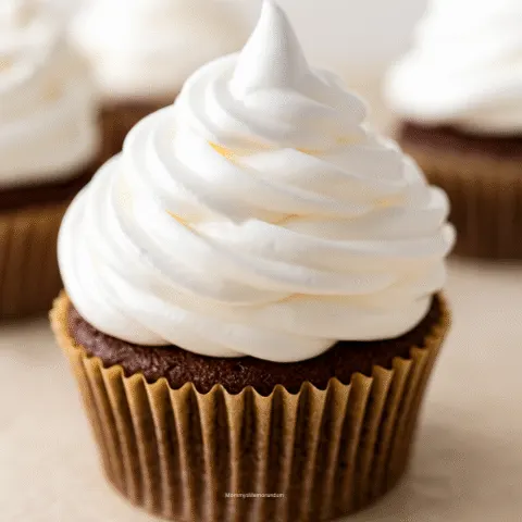Close-up of chocolate cupcake topped with glossy, swirled marshmallow frosting. Background includes additional cupcakes with similar frosting, styled on a neutral surface.