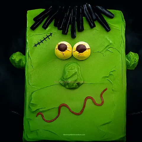Frankenstein-themed Halloween brownie with green frosting, candy eyes, black licorice hair, and red licorice mouth, set against a spooky dark background with fog-like texture.