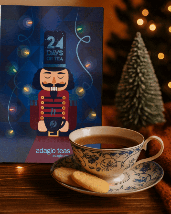 Cozy holiday tea scene with Adagio Teas &lsquo;24 Days of Tea&rsquo; Advent Calendar, creamy white teacup with blue-and-silver pattern, tea cookies on the saucer, warm twinkle lights, and a small frosted Christmas tree &mdash; festive tea setup for holiday gift inspiration