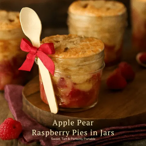 Four Apple Pear Raspberry Pies baked in mason jars with golden crusts and sugar sprinkles, styled on a rustic wooden board with a red ribbon and wooden spoon tied around one jar. Warm-toned background with raspberries and a burgundy napkin.