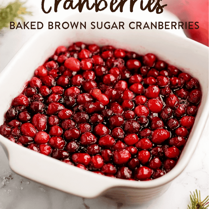 Close-up image of baked brown sugar cranberries in a white ceramic baking dish, styled with festive holiday props like pine sprigs and a red napkin. Cranberries are glossy, caramelized, and glistening under warm lighting — perfect for a cozy Thanksgiving or Christmas side dish.