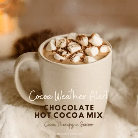 A steaming mug of hot cocoa topped with marshmallows and chocolate shavings, set on a chunky knit blanket with warm candlelight and pinecones in the background.