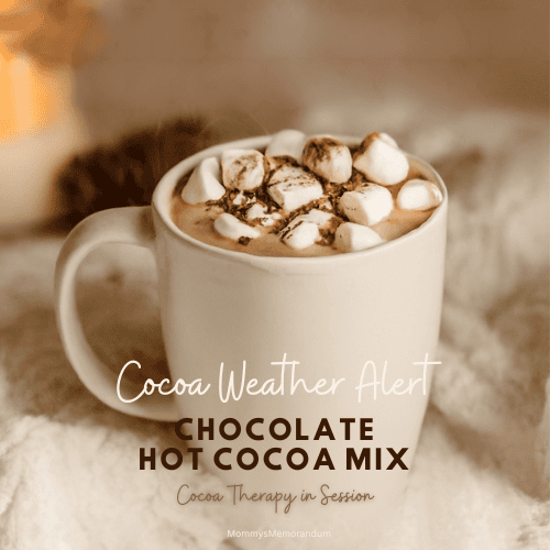 A steaming mug of hot cocoa topped with marshmallows and chocolate shavings, set on a chunky knit blanket with warm candlelight and pinecones in the background.