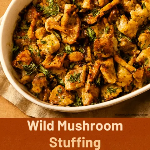 Overhead view of wild mushroom stuffing in a white ceramic dish, filled with toasted ciabatta cubes, chanterelle and blue foot mushrooms, and fresh herbs. Cozy holiday table setting with warm tones and autumn decor.