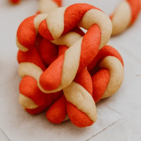 Close-up of twisted red and white candy cane cookies stacked on parchment paper. The cookies have a festive swirl and curved tops, styled for the holidays with soft lighting and shallow depth of field.