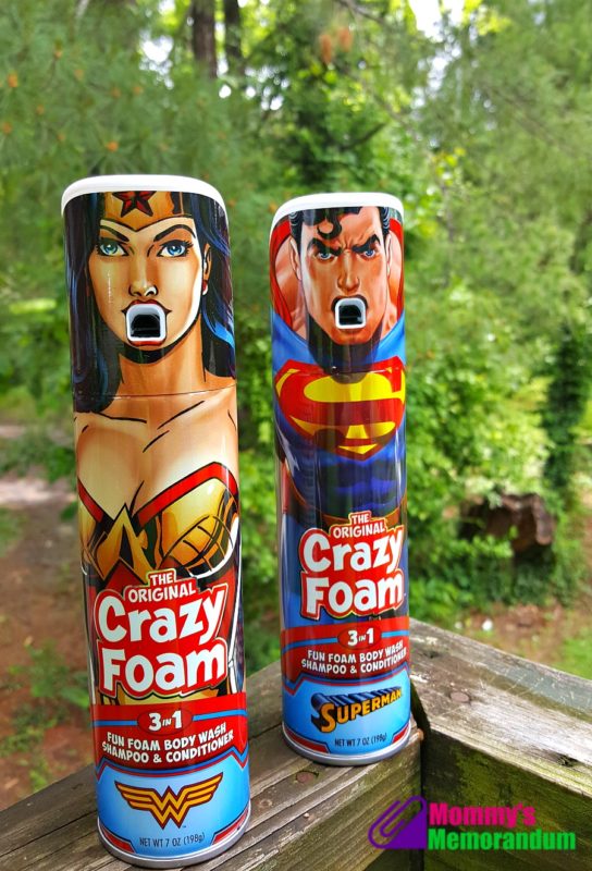 Two cans of Crazy Foam featuring Wonder Woman and Superman designs, placed on a wooden railing with a greenery background.