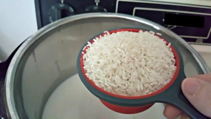 A close-up photo of a hand pouring measured rice into the instant pot bowl whch contains milk, sugar, and salt for Instant Pot rice pudding.