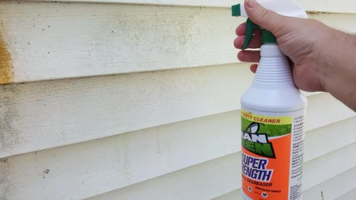 Hand holding a bottle of Mean Green Super Strength Cleaner, spraying it onto dirty white house siding.