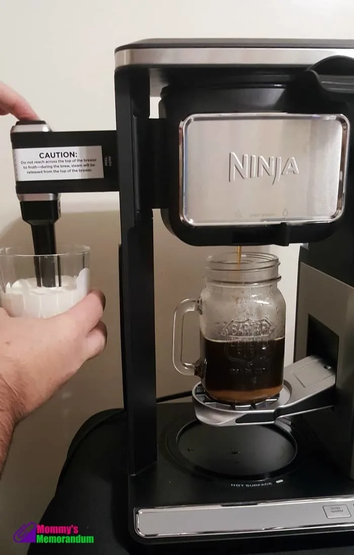 A Ninja coffee machine brewing toffee coffee into a glass mug, while a hand prepares to froth milk in a separate glass.