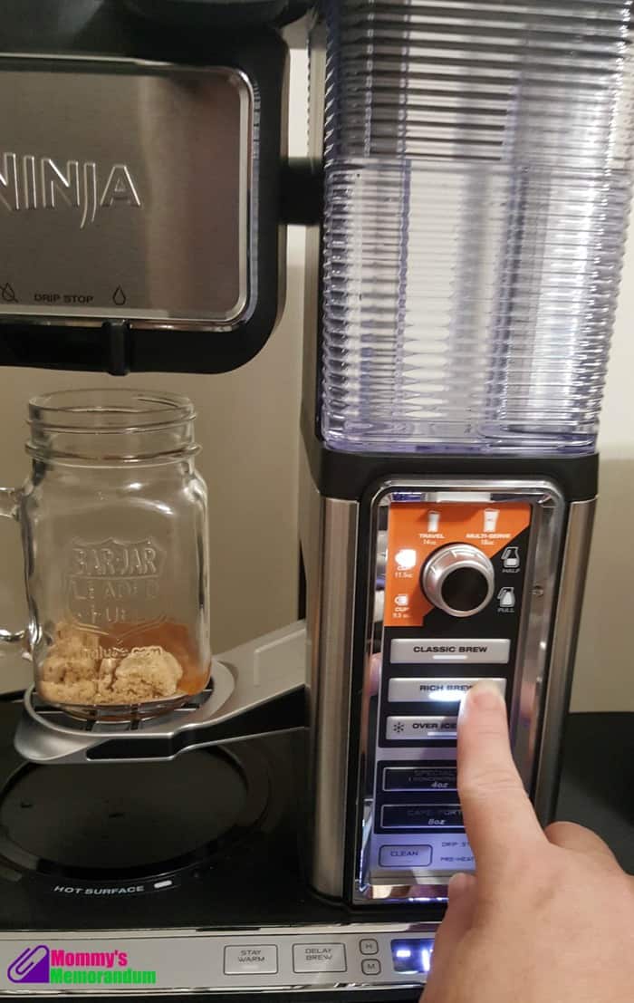 A hand pressing the Rich Brew button on a Ninja coffee machine, with a glass mug containing toffee coffee ingredients.